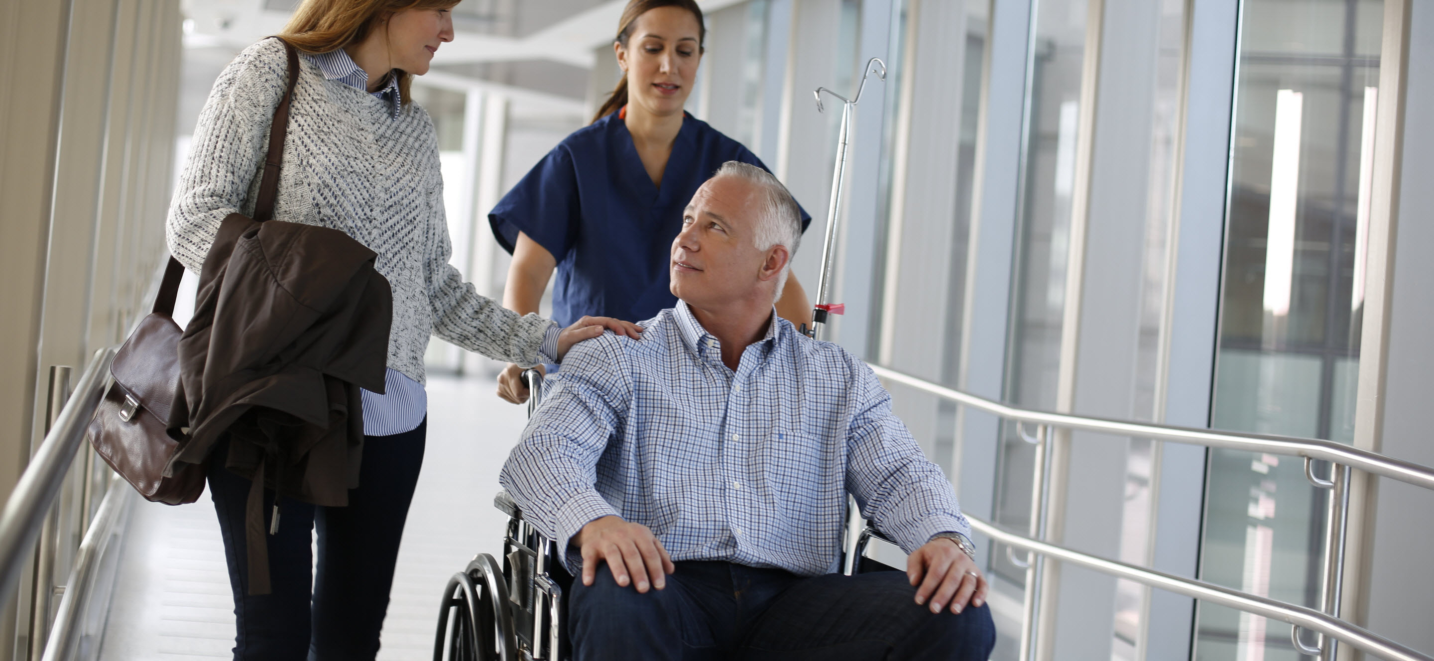Older man in wheelchair looks up at daughter as healthcare professional pushes the wheelchair.