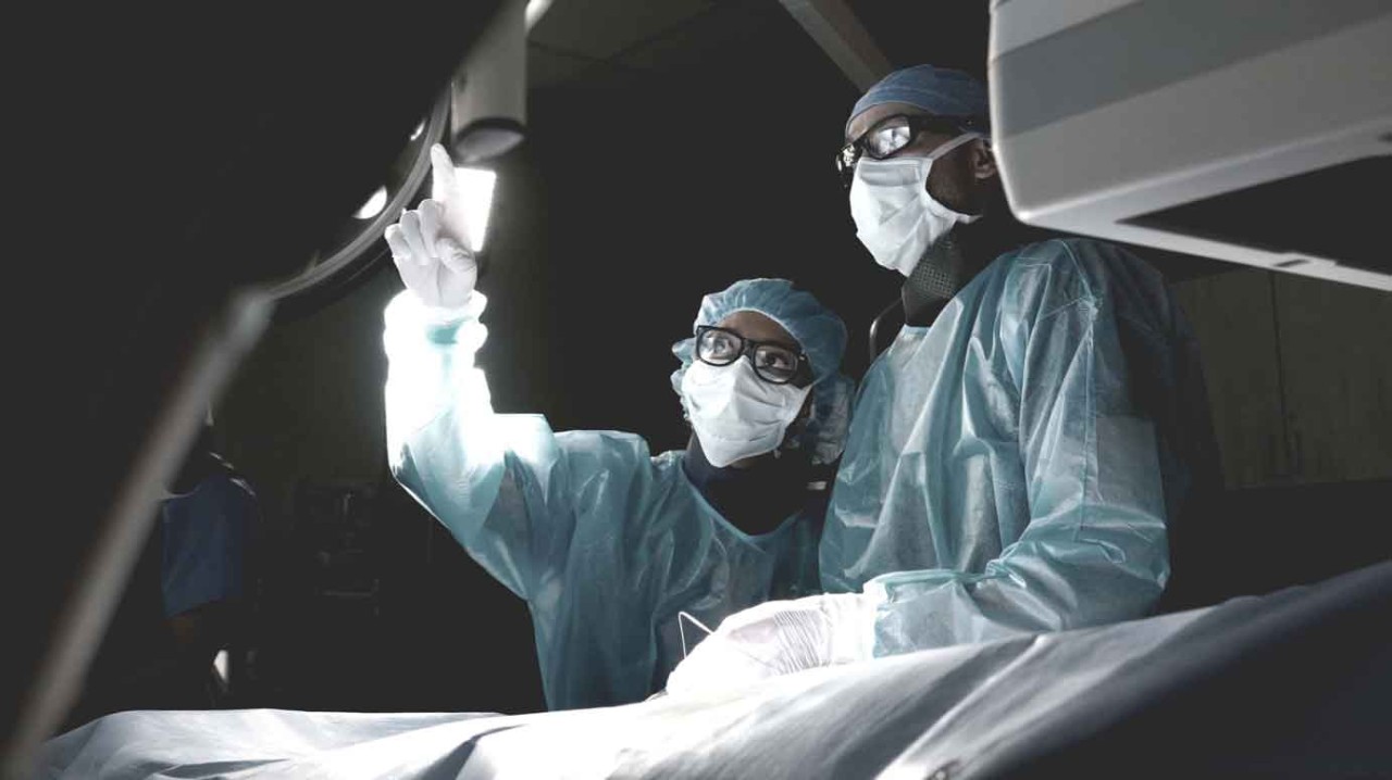 Two healthcare professionals in operating room and surgical gear looking up as one points.